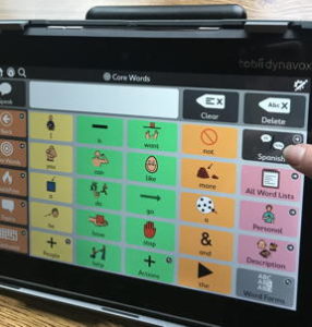 A 6 by 5 English speech generating device grid is displayed. A finger is pointing to a button that reads "Spanish."